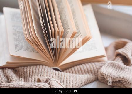 Warmes und gemütliches Hygge-Konzept mit weißem und beigefarbenem Pullover, altes geöffnetes Buch auf Fensterbank, getönt Stockfoto
