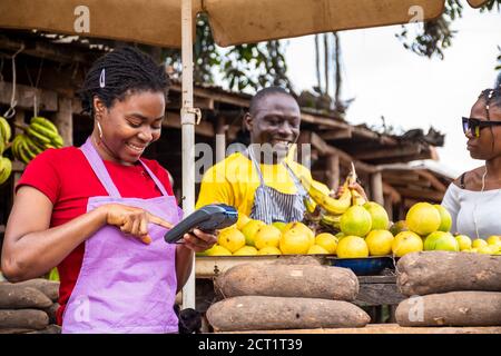 Junge Frau mit einer POS-Maschine in einem lokalen Markt, Mann verkauft eine Kochbanane für eine Dame im Hintergrund Stockfoto