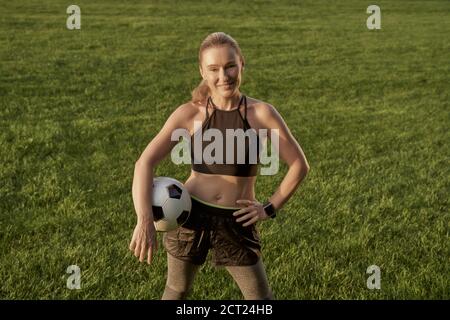 Fußballspieler. Schöne glückliche Frau hält Fußball und lächelt an der Kamera, während sie im Freien in einem grünen Feld steht. Fußball spielen Stockfoto