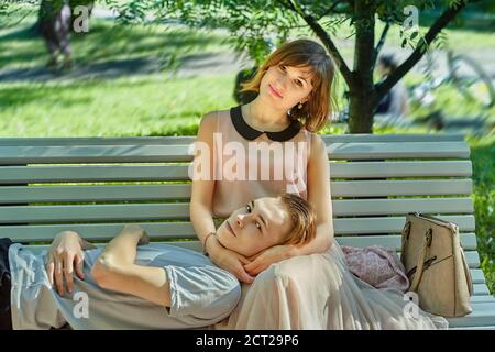 Junge schöne Frau sitzt auf der Bank mit dem Kopf ihres Freundes auf den Knien. Kaukasisches Paar hat ein Date im öffentlichen Park. Stockfoto