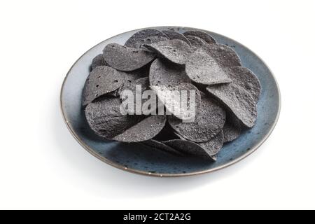 Schwarze Kartoffelchips mit Holzkohle auf einer blauen Keramikplatte isoliert auf weißem Hintergrund. Seitenansicht, Nahaufnahme. Stockfoto
