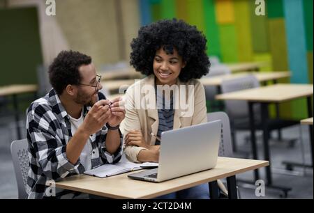 Zwei junge, glückliche Mixed-Race-Kollegen sitzen am Schreibtisch im Coworking Space, diskutieren mit einem Laptop über ein neues Projekt, der lächelnden Kollegin etwas erklärt. Geschäftsleute, Teamarbeit Stockfoto