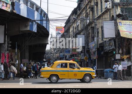 Kalkutta, Indien - 1. Februar 2020: Unbekannte Menschen stehen und gehen auf der Straße, während ein traditionelles gelbes Taxi unter Maa Flyover vorbei fährt Stockfoto