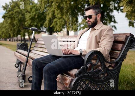 Bärtiger junger Geschäftsmann in eleganter Casualwear und Sonnenbrillen beim Stöbern Das Netz Stockfoto
