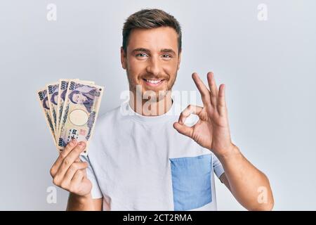 Schöner kaukasischer Mann hält 5000 japanische Yen-Banknoten tun ok Zeichen mit Fingern, lächelnd freundlich gestikulieren ausgezeichnetes Symbol Stockfoto