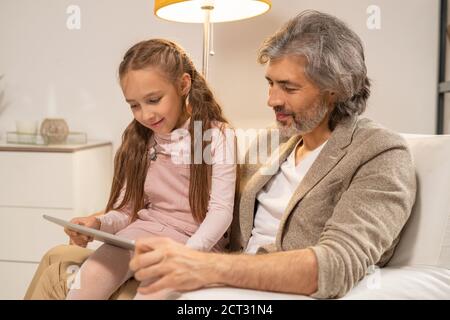 Glücklicher Großvater hält seine Enkelin auf den Knien, während er im Sitzen sitzt Sessel Stockfoto