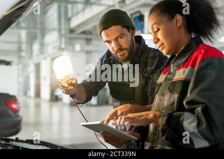 Junge bärtige Mann in Arbeitskleidung zeigt auf Tablet-Display gehalten Von seinem Kollegen Stockfoto