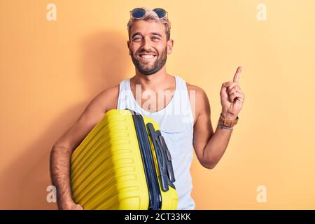 Junge schöne blonde Tourist Mann im Urlaub trägt Sonnenbrille halten Flugzeugkabine Tasche lächelnd glücklich zeigen mit Hand und Finger Zur Seite Stockfoto