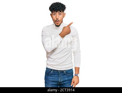 Junger arabischer Mann trägt lässigen Winter Pullover überrascht zeigt mit Finger zur Seite, offenen Mund erstaunt Ausdruck. Stockfoto