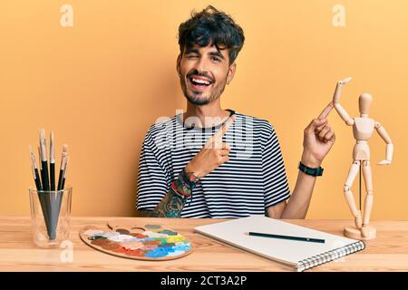 Junger hispanischer Mann sitzt lächelnd auf dem Tisch im Kunststudio und schaut auf die Kamera, die mit zwei Händen und Fingern zur Seite zeigt. Stockfoto