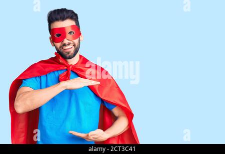 Junger schöner Mann mit Bart trägt Superhelden Kostüm gestikulieren mit Händen zeigt große und große Größe Zeichen, Maßnahme Symbol. Lächelnd Blick auf die Stockfoto