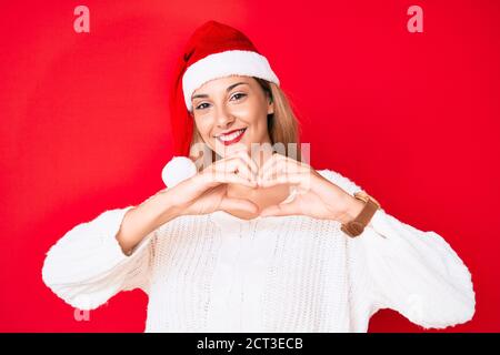 Junge Brünette Frau trägt weihnachtshut lächelnd in der Liebe zeigt Herz Symbol und Form mit Händen. Romantisches Konzept. Stockfoto