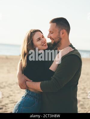Junges erwachsenes Paar, das an der Küste steht und sich anschaut. Schöner lächelnder Mann umarmt schöne Frau voller Liebe. Millennial männlich und weiblich posiert am Strand an sonnigen Tag Stockfoto