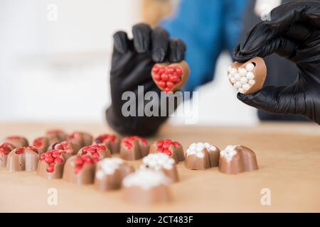 Konditor in schwarzen Handschuhen macht süße herzförmige Pralinen mit rot-weißen Füllung Stockfoto