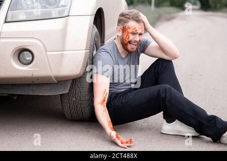 Der junge Mann sitzt mit blutigem Kopf in der Nähe des Wagens, schreiend und bereutend handeln Stockfoto