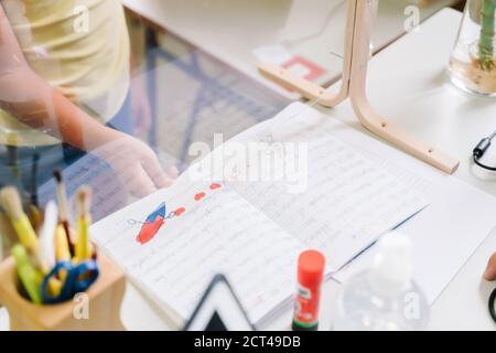 Schüler mit einer Maske, die Hausaufgaben an den Lehrer übergibt Durch einen Bildschirm Stockfoto