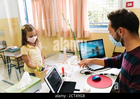 Schüler mit einer Maske, die Hausaufgaben an den Lehrer übergibt Durch einen Bildschirm Stockfoto