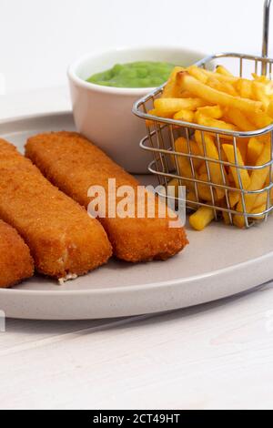 Fischfinger mit Chips in einem Drahtkorb und matschigen Erbsen serviert. Weißer Holzhintergrund Stockfoto