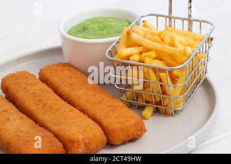 Fischfinger mit Chips in einem Drahtkorb und matschigen Erbsen serviert. Weißer Holzhintergrund Stockfoto
