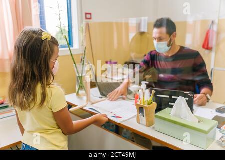 Schüler mit einer Maske, die Hausaufgaben an den Lehrer übergibt Durch einen Bildschirm Stockfoto