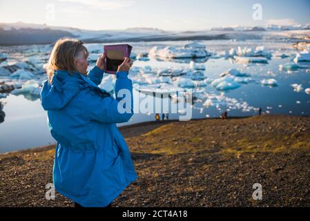 Frau, die mit einem ipad ein Foto im Urlaub an der Jokulsarlon Glacier Lagoon macht, einem Gletschersee voller Eisberge in Südostisland, Europa Stockfoto