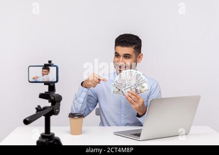 Finanzielle Bildung. Glücklicher Geschäftsmann sitzt am Arbeitsplatz, zeigt Dollar-Banknoten und lächelt aufgeregt, Aufnahme natürlich, wie Geld verdienen Gewinn. Stockfoto