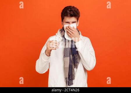 Traurige ungesunde junge Mann in lässigen Pullover mit Kapuze und karierten Schal am Hals hält Tablette in der Hand, leiden an Grippe, laufende Nase. Innen Stockfoto