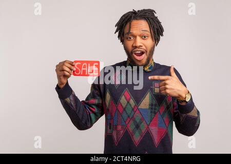 Überrascht afrikanischen Mann mit Dreadlocks schockiert mit niedrigen Kosten Zeigefinger auf Verkauf Zeichen in der Hand mit offenem Mund und großen Augen. Innenaufnahme im Studio Stockfoto