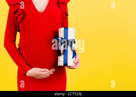 Zugeschnittenes Bild von schwangeren Frau in rotem Kleid mit einer Geschenkbox und berühren ihren Bauch auf gelbem Hintergrund. Erwartet einen kleinen Jungen. Speicherplatz kopieren. Stockfoto