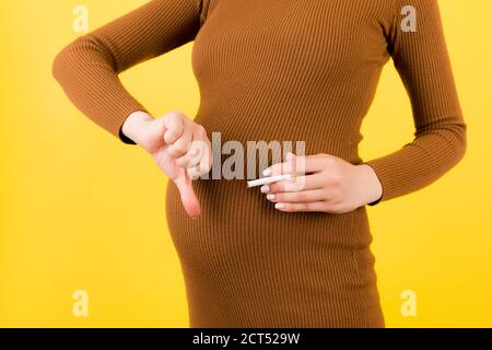 In der Nähe von schwangeren Frau in braunem Kleid hält eine Packung Zigaretten und zeigt, dass ich es nicht mag Geste auf gelbem Hintergrund. Daumen nach unten. Schlechter Habbi Stockfoto