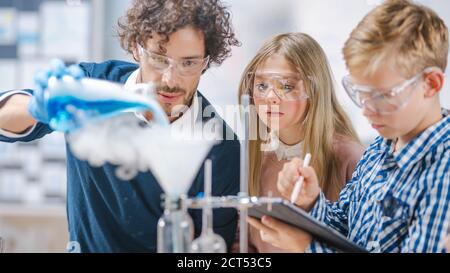 Grundschule Wissenschaft Klassenzimmer: Begeisterter Lehrer erklärt Chemie zu verschiedenen Gruppe von Kindern, zeigt ihnen, wie man Chemikalien in Bechern mischen Stockfoto