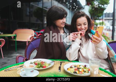 Die reife Mutter und ihre junge Tochter sitzen zusammen im Café oder Restaurant. Frauen mit guten Beziehungen Blick auf gelbe Smartphone und lachen Stockfoto