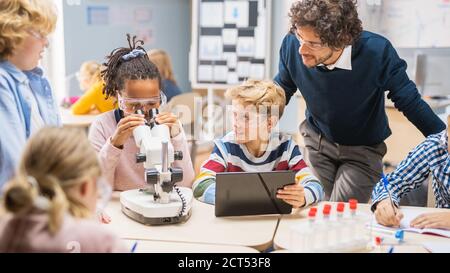 Elementary School Science Classroom: Cute Little Girl Looks under Microscope, Boy nutzt Digital Tablet Computer, um Informationen im Internet zu überprüfen Stockfoto