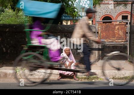 Eine ältere Dame sitzt allein vor ihrem Pflegeheim, auf einer belebten Straße, während eine Rikscha in Delhi, Indien, vorbei rast Stockfoto