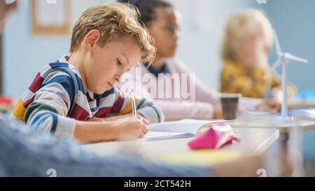 In der Grundschule Klassenzimmer Brilliant Kaukasischen Jungen schreibt in Übung Notebook, die Prüfung und Schreiben Prüfung. Junior-Klassenzimmer mit Gruppe von Stockfoto