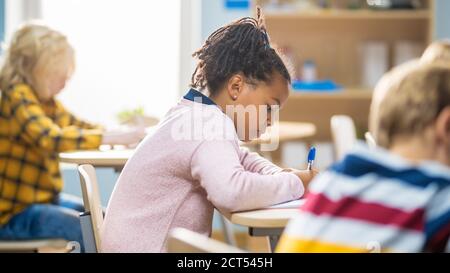 In Grundschule Klassenzimmer Brilliant Black Girl schreibt in Übung Notebook, die Prüfung und Schreiben Prüfung. Junior-Klassenzimmer mit Gruppe von hellen Stockfoto