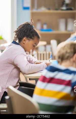 In Grundschule Klassenzimmer Brilliant Black Girl schreibt in Übung Notebook, die Prüfung und Schreiben Prüfung. Junior-Klassenzimmer mit Gruppe von hellen Stockfoto