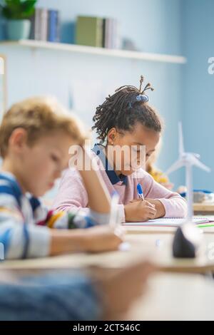 In Grundschule Klassenzimmer Brilliant Black Girl schreibt in Übung Notebook, die Prüfung und Schreiben Prüfung. Junior-Klassenzimmer mit Gruppe von hellen Stockfoto