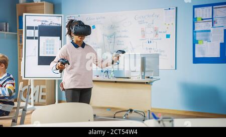 Grundschule Informatik-Klasse: Nettes Mädchen trägt Augmented Reality-Headset und mit Controllern lernt Lektionen in Virtual Reality aufgeregt Stockfoto