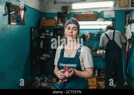 Gleichstellung der Geschlechter. Porträt einer jungen lächelnden Frau in Uniform, die in einer Werkstatt arbeitet, die sich mit einem Lappen die Hände wischt. Im Hintergrund ein Arbeiter bei Th Stockfoto