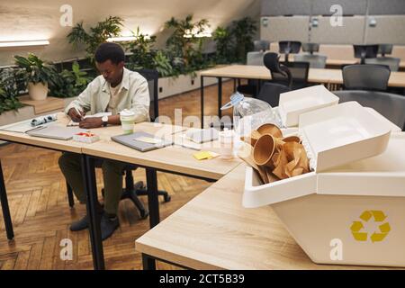Hintergrundbild von zwei Abfallsortiercontainern auf dem Schreibtisch in moderner Büroeinrichtung mit afroamerikanischem jungen Mann, der arbeitet, Kopierraum Stockfoto