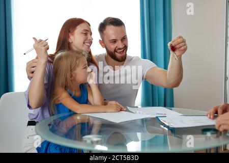 Glücklicher Mann mit Frau und Tochter bekommen Schlüssel aus ihrem ersten Haus, sitzen sie aufgeregt, unterschreiben die Dokumente, um Hypothek zu bekommen Stockfoto