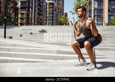 Junger Mann, der draußen trainiert. Guy hält Gummiband auf den Schultern und hocken Übungen. Hartes intensives Training oder Workout. Sport im Freien Stockfoto