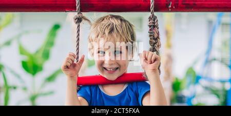 Portrait schließen Happy Smile Sommersprosse blauäugig niedlicher blonder kleiner Junge sitzt auf einer Seilleiter und spielt draußen auf dem Spielplatz im Kindergarten. Kindertagesstätte Stockfoto