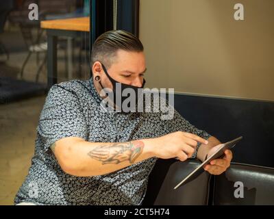 Aufnahme eines Mannes in einer medizinischen Maske in einem Cafe schaut auf sein Tablet - neu normal Stockfoto