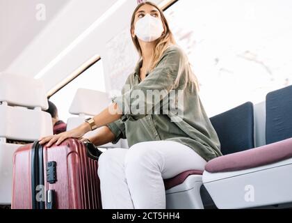 Von unten junge Frau in Schutzmaske berühren Koffer und Wegschauen, während auf dem Sitz im modernen Zug sitzen während Urlaub Stockfoto