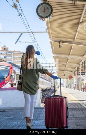 Rückansicht der unkenntlich Frau in Latexhandschuhen ziehen Koffer Und die Zeitkontrolle beim Gehen auf Bahnsteig des Bahnhofs Während der Epidemie Stockfoto