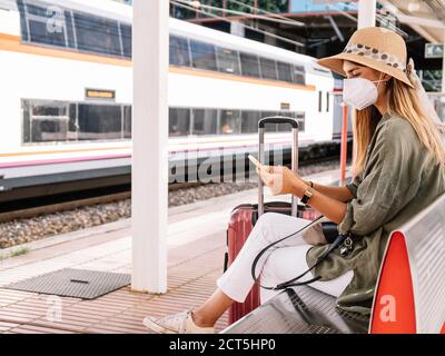 Seitenansicht der jungen Frau in Schutzmaske sitzend auf Bank auf Plattform des Bahnhofs und mit Smartphone während Sommerurlaub Stockfoto