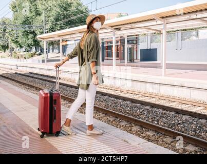 Seitenansicht der jungen Frau in medizinischer Maske und mit Koffer steht auf Plattform und wartet auf Zug auf der Bahn Station während einer Pandemie Stockfoto