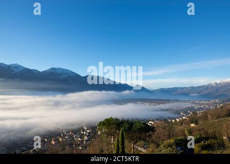 Alpine Village Locarno unter Nebelmeer über einem Alpensee Maffiore mit schneebedeckter Berg im Tessin, Schweiz. Stockfoto